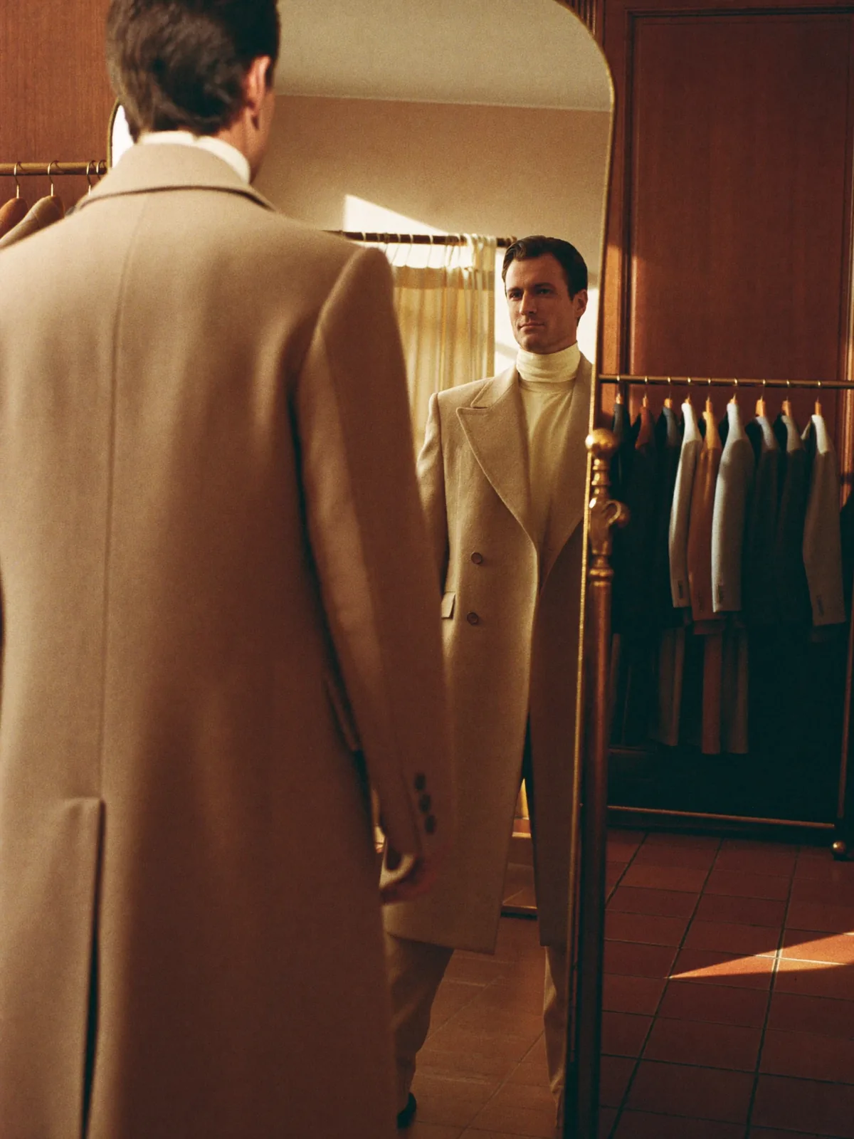 A man in a camel overcoat over a cream roll-neck studying himself in a boutique fitting mirror.